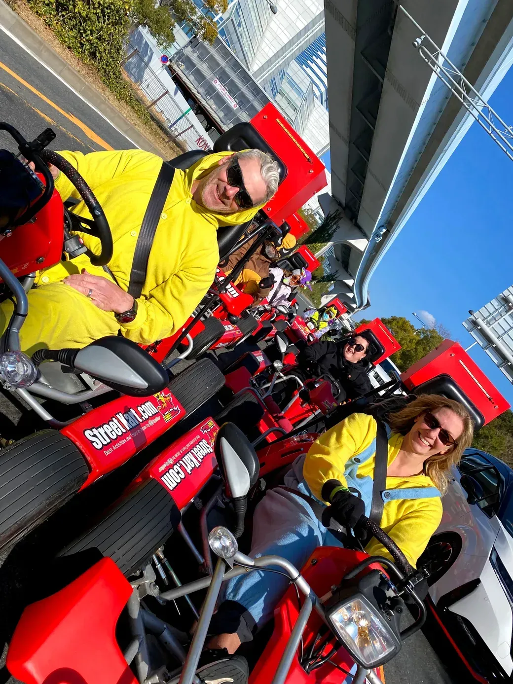 Group of people in yellow jackets riding red promotional trikes on a city street, smiling at the camera.