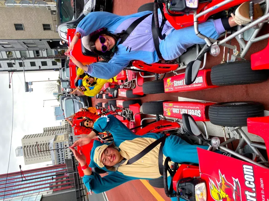 Group of people in bright red go-karts lined up on a city street, smiling and waving at the camera.