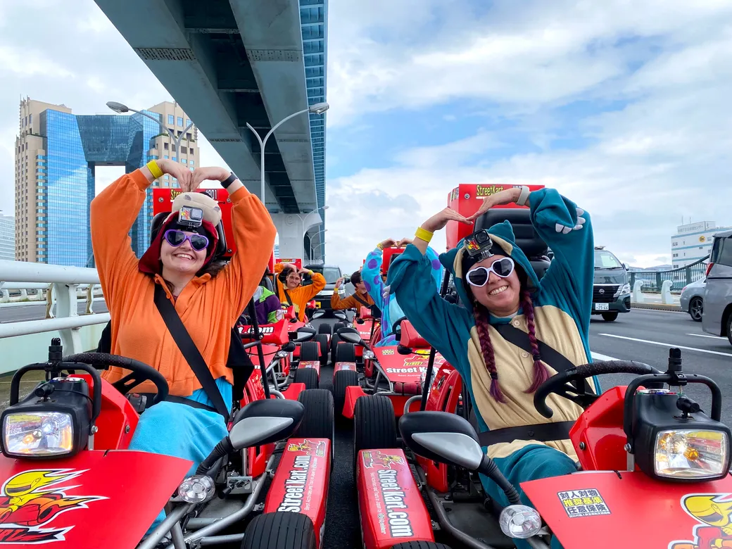 Group of people in colorful outfits on go-karts on a bridge, forming heart shapes with their arms and smiling at the camera.