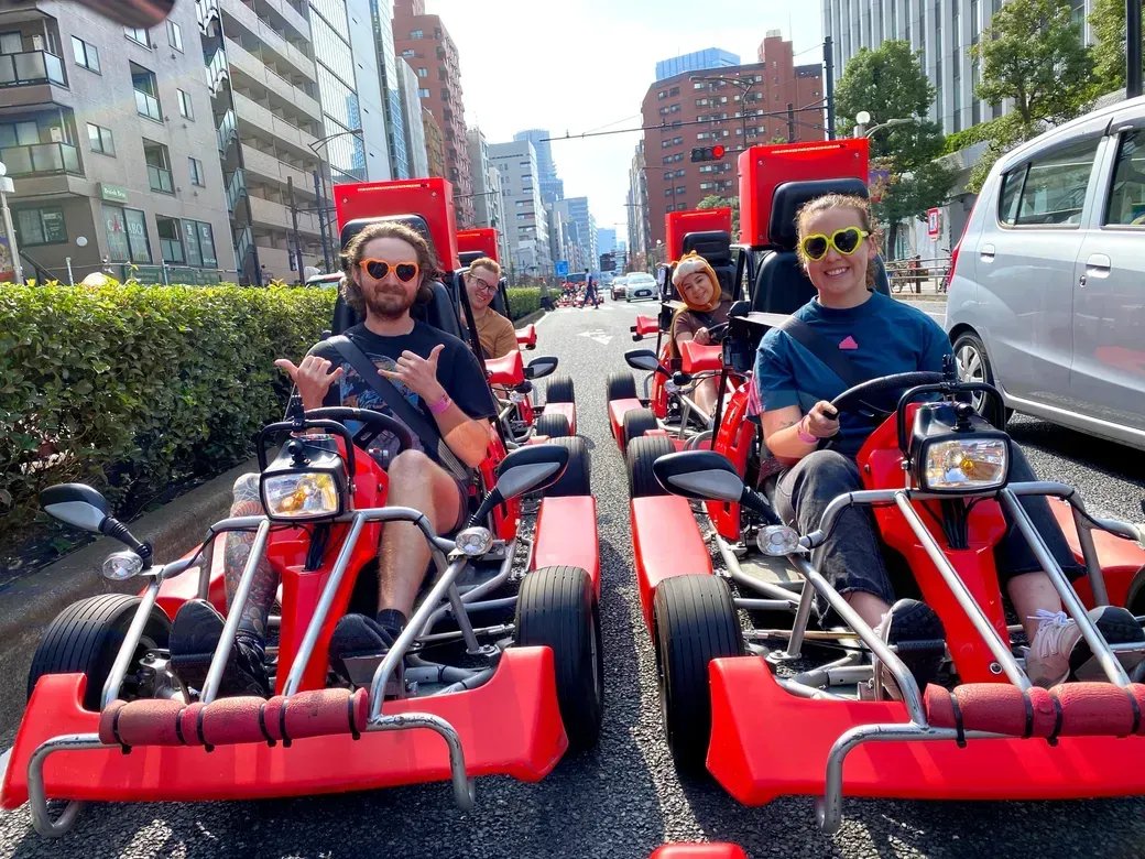 A group of friends riding bright red pedal go-karts down a city street, smiling and wearing sunglasses.